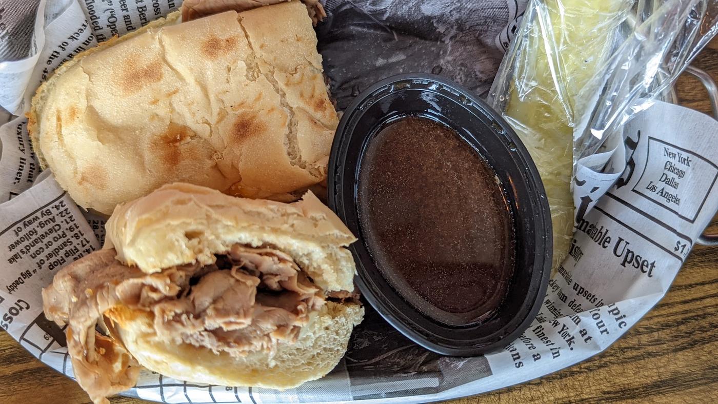 A shredded meat sandwich on a hoagie roll in a newsprint lined basket, with a cup of jus and a wrapped pickle spear.
