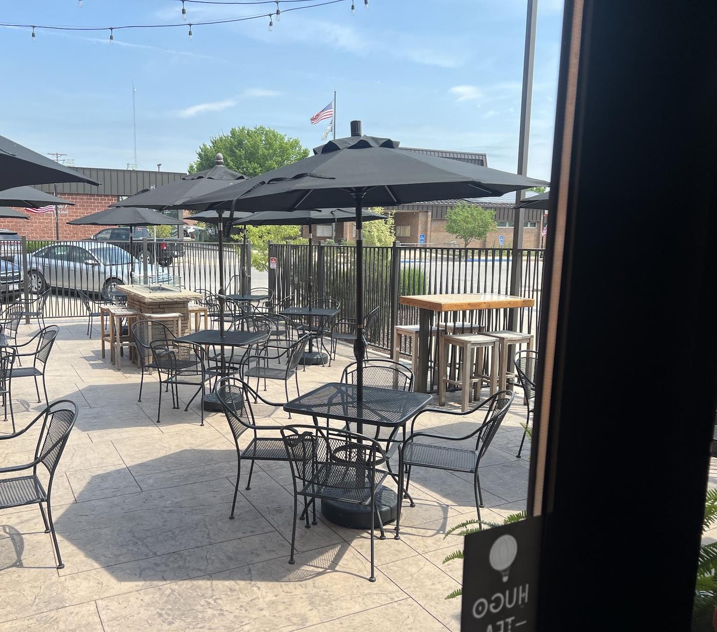 The patio: black metal tables and chairs under gray umbrellas, string lights overhead, a stone fire pit table and a wrought iron railing.